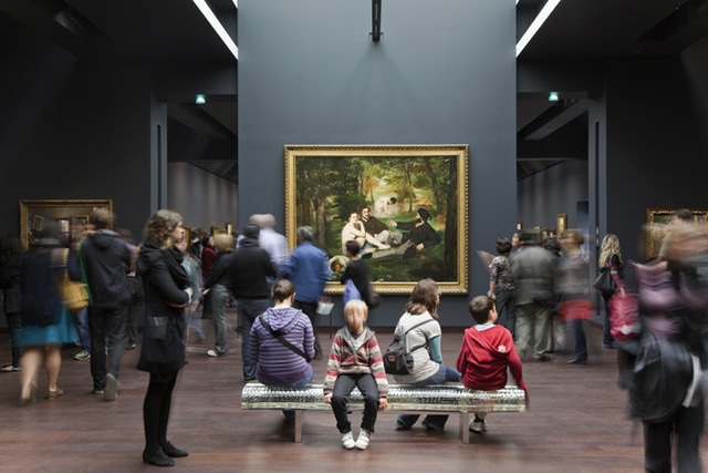 Photo of people watching master pieces in Orsay's Impressionist Gallery (in the background Luncheon on the Grass by Édouard Manet), to illustrate an Orsay Museum Guided Tour in Paris, France.